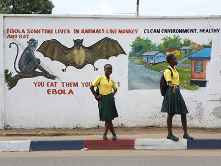 Two school girls stand on the street in front of a wall with a health message about Ebola