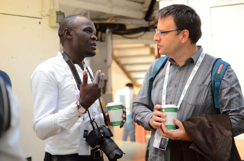 Issa, with a camera hanging around his neck, speaks with a man holding a cup 