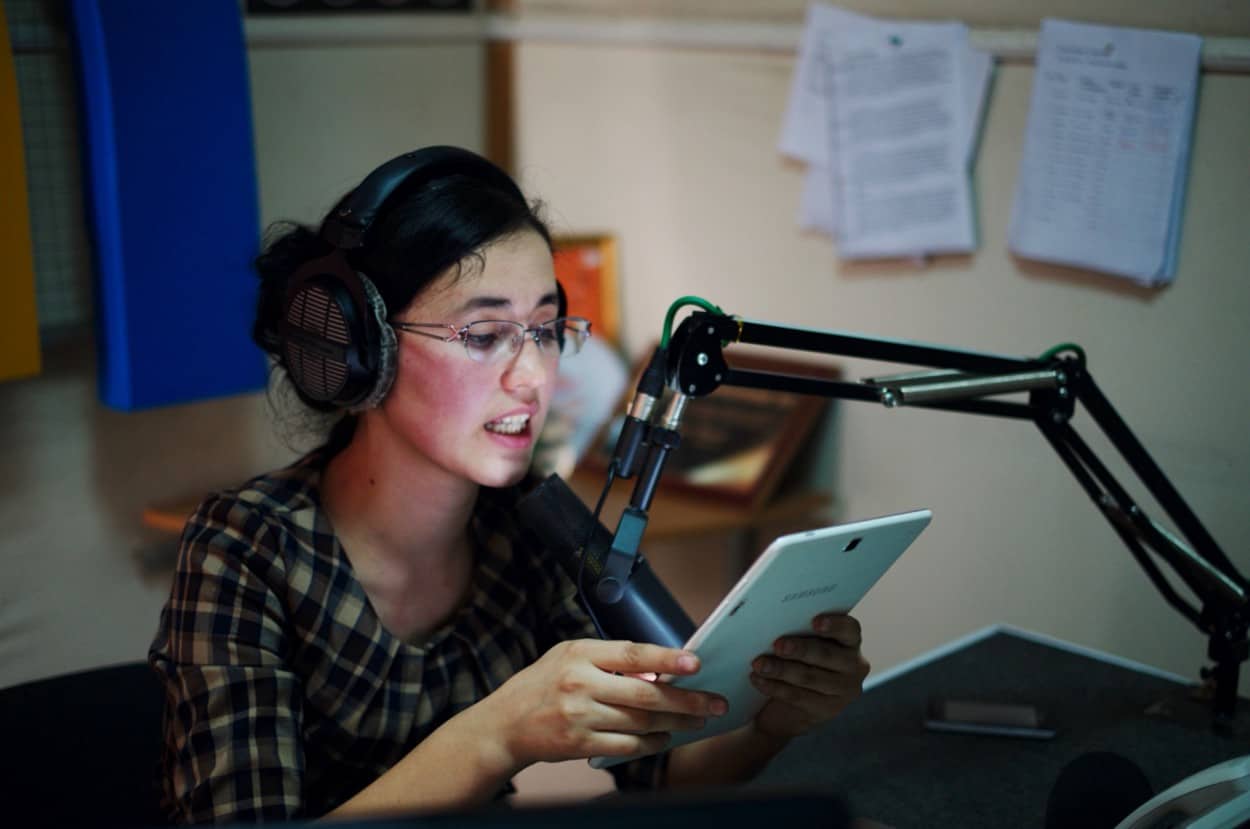 A woman wearing headphones sits at a mic reading from a tablet.