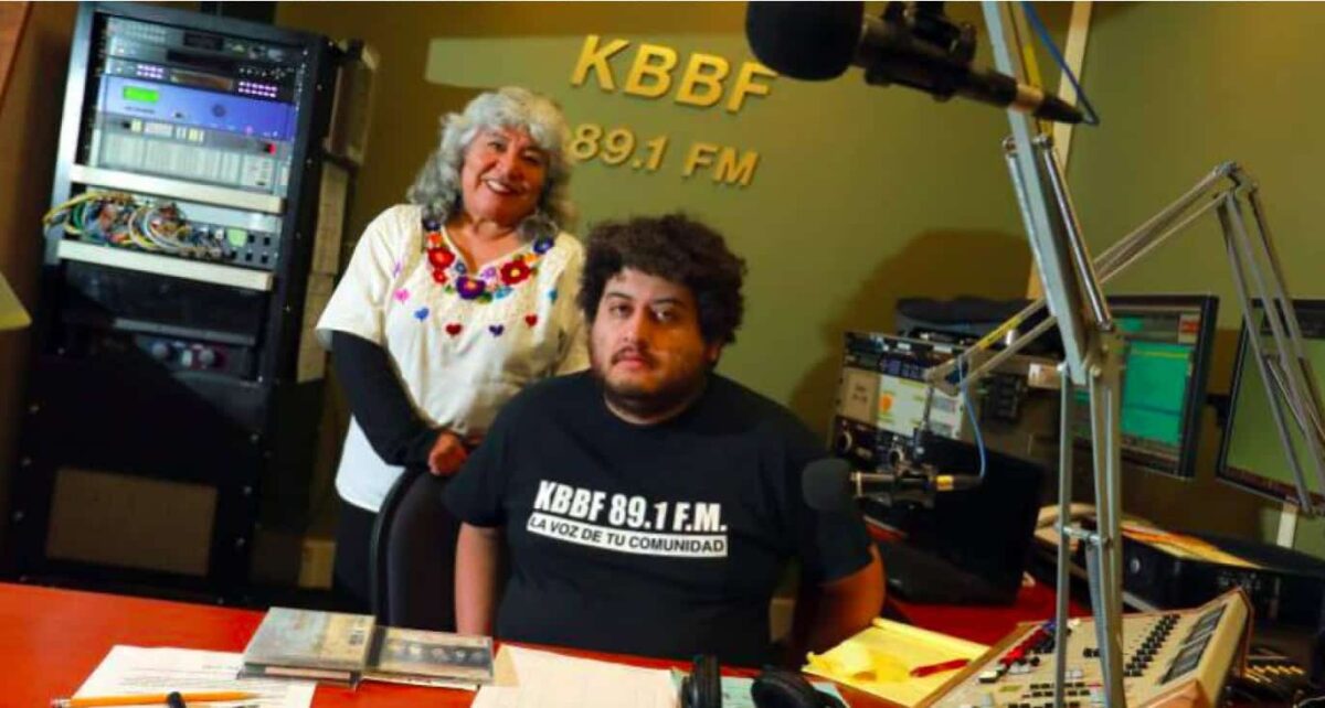 A man sits at a table with an older woman standing behind him. "KBBF 89.1 FM" is on the wall behind them.