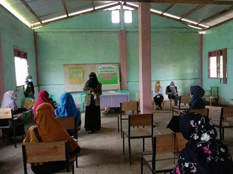 Inside a building, a group of young women sit spaced out; one woman is standing