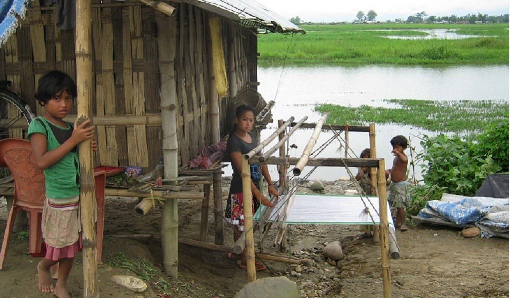 Children stand outside a shack made of bamboo next to a river