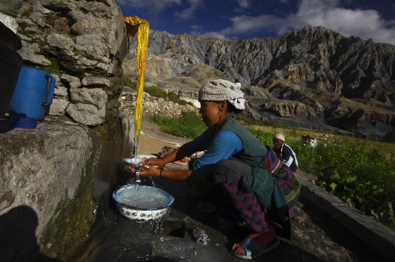 A woman washes dishes at a water fountain in the mountains