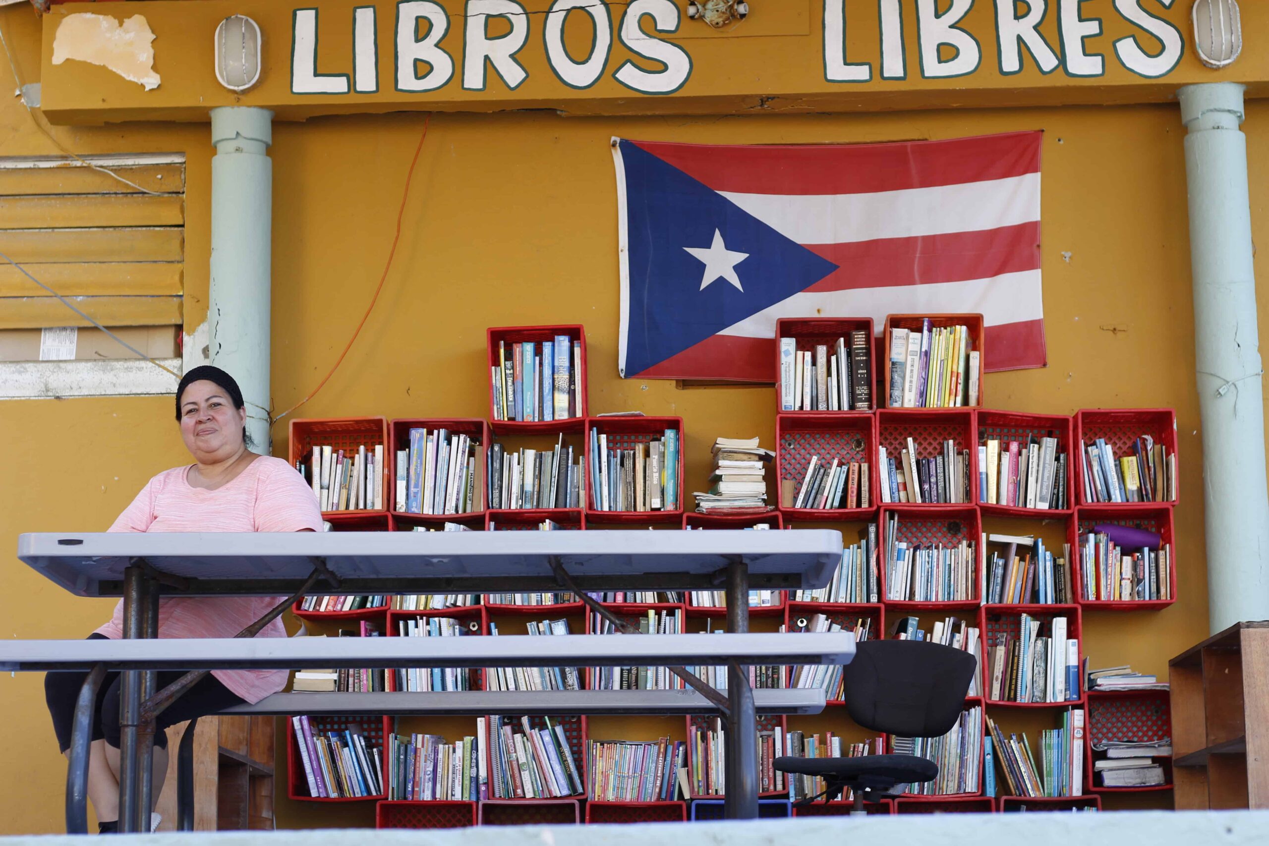 A woman sits at a picnic table in front of bookshelves filled with books and a Puerto Rican flag.