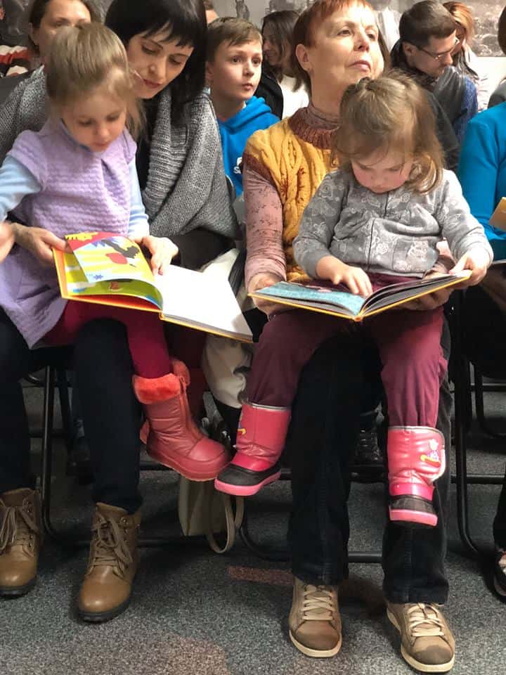 Two girls holding books sit on their mothers' laps