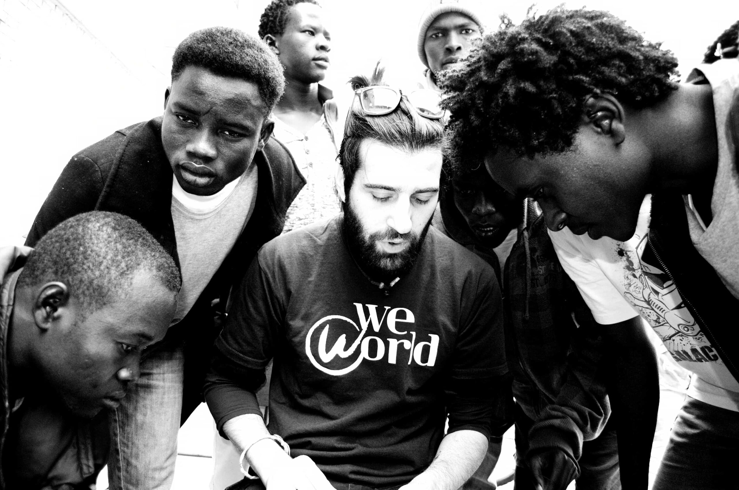 A white man with a beard sits. A group of young black men stand around him.
