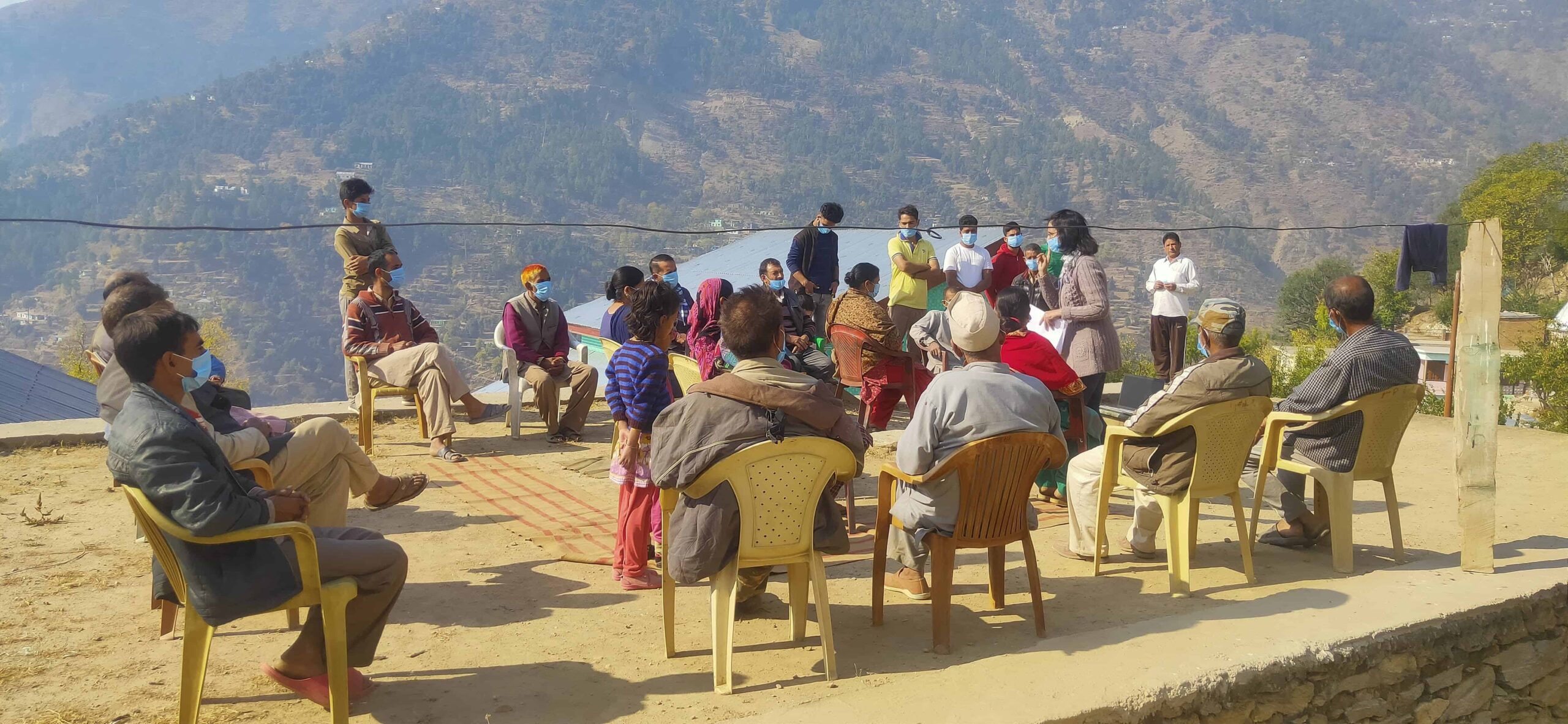 A group of people wearing masks sits on plastic chairs outside on a concrete pad. A woman wearing a mask stands talking to them.