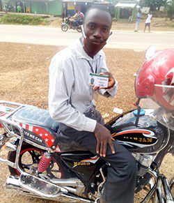 A man sits on a motorbike wearing a face mask and holding a card