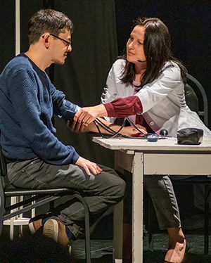 A woman and man sit at a small table while the woman takes the man's blood pressure