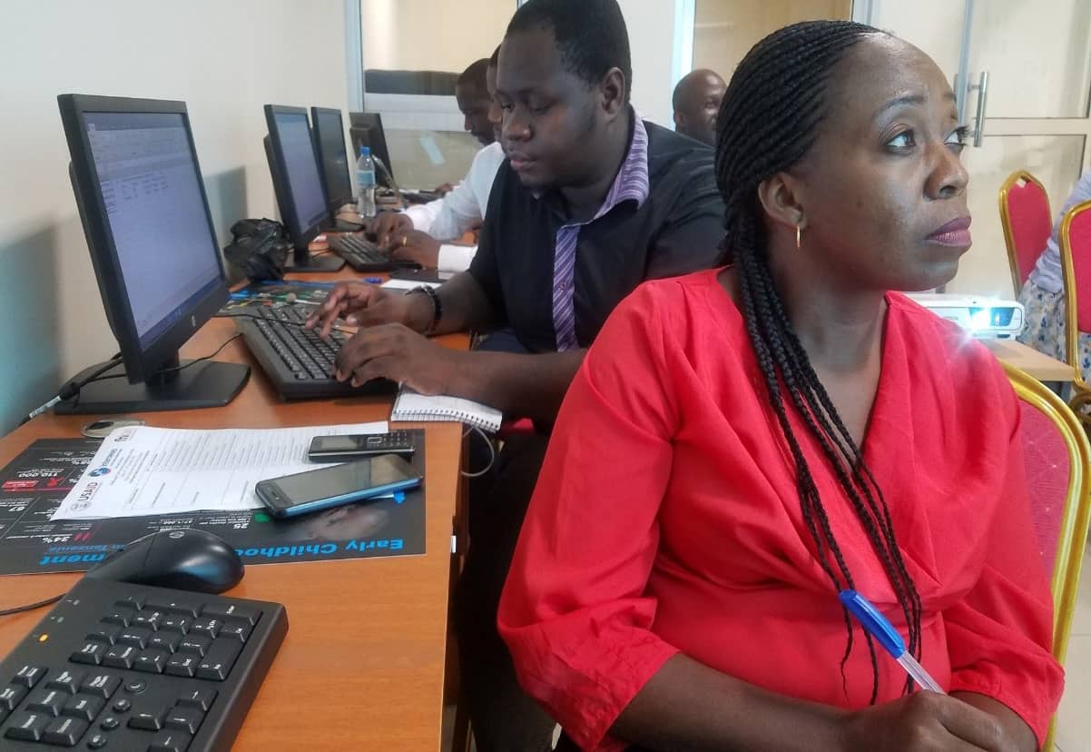 A woman sits by a table with computer monitors on it. A man sits behind her.