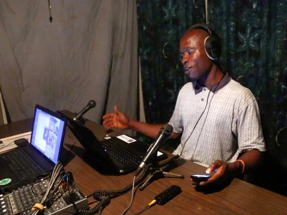 A man wearing headphones sits in front of a laptop and a microphone