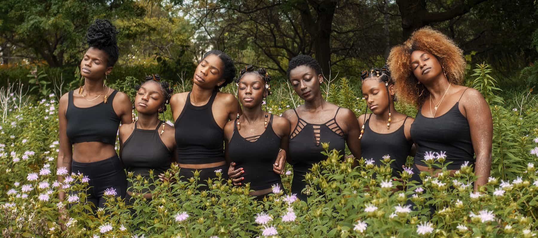 A group of seven Black women and girls stand in a field of flowers; all are dressed in black and have their eyes closed