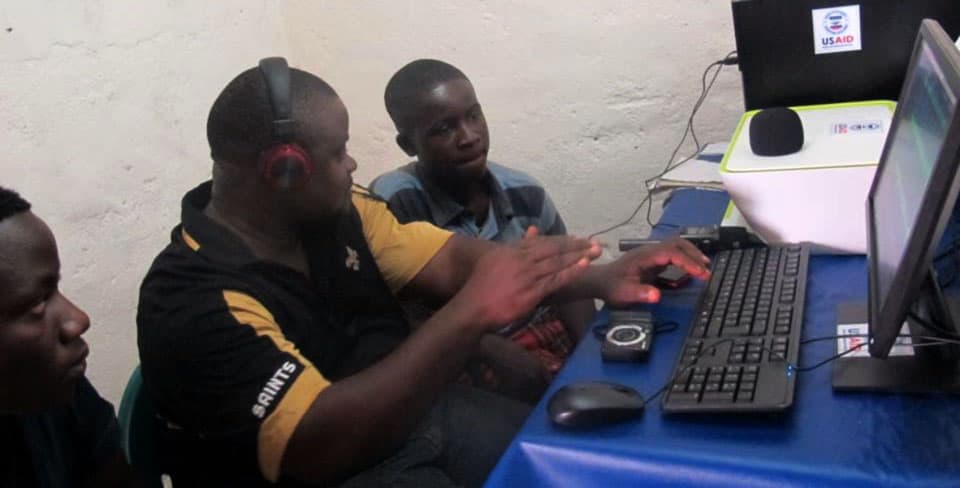 A man wearing headphones sits at a computer; there are two young men sitting on either side.