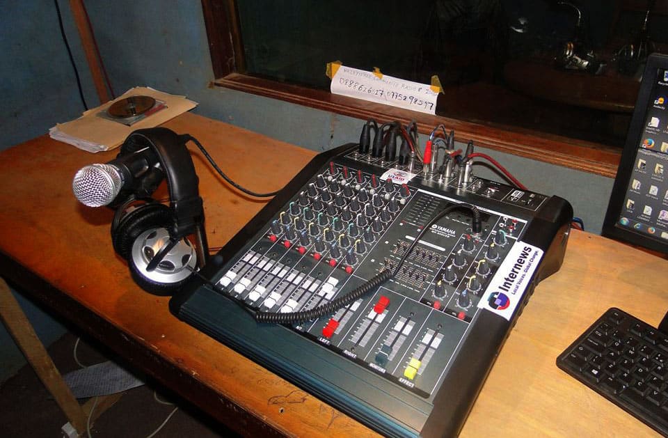 A mixing board, headphones and a mic sit on a weathered table in a radio studio