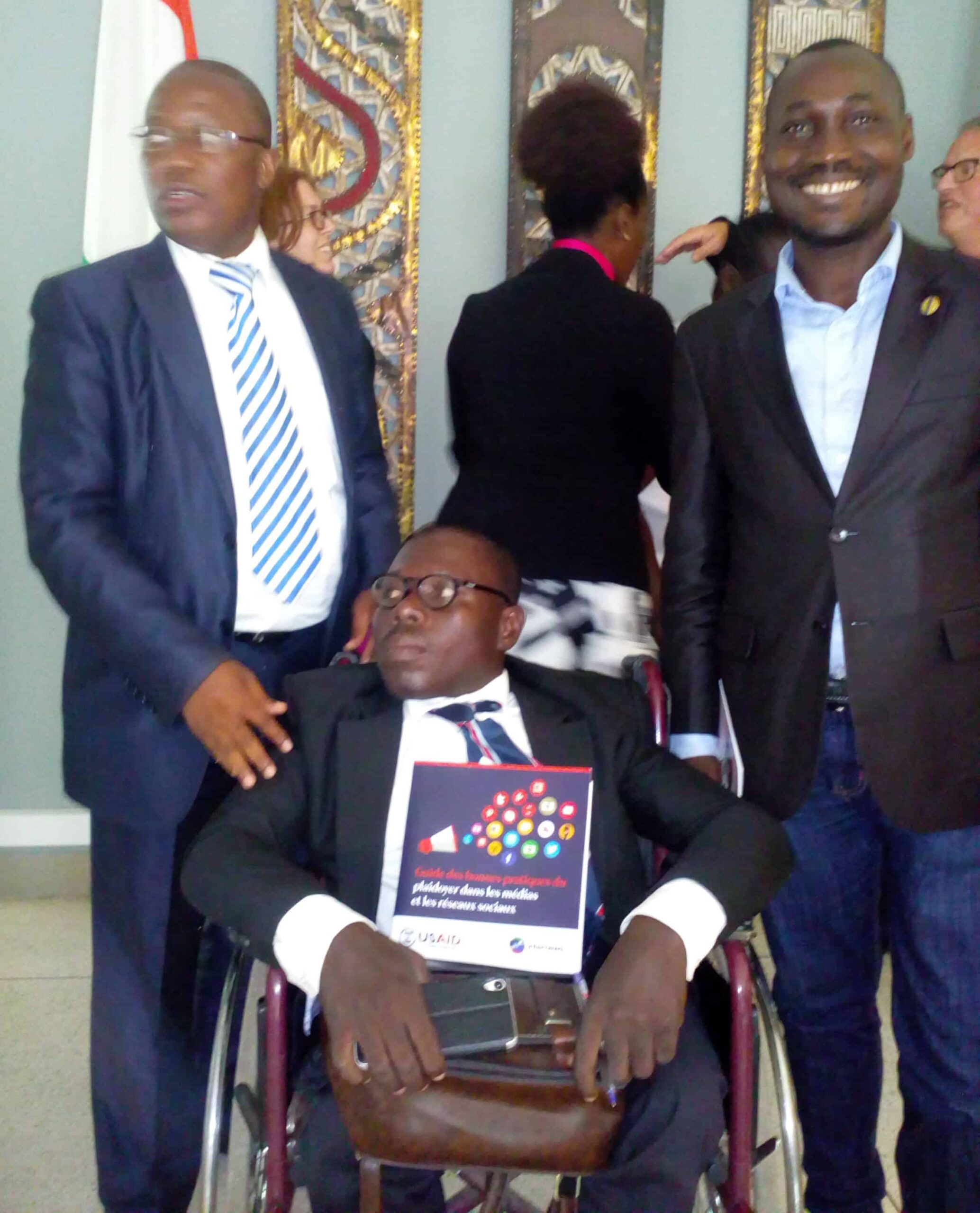 Two men stand next to a man in a wheelchair who is holding a book