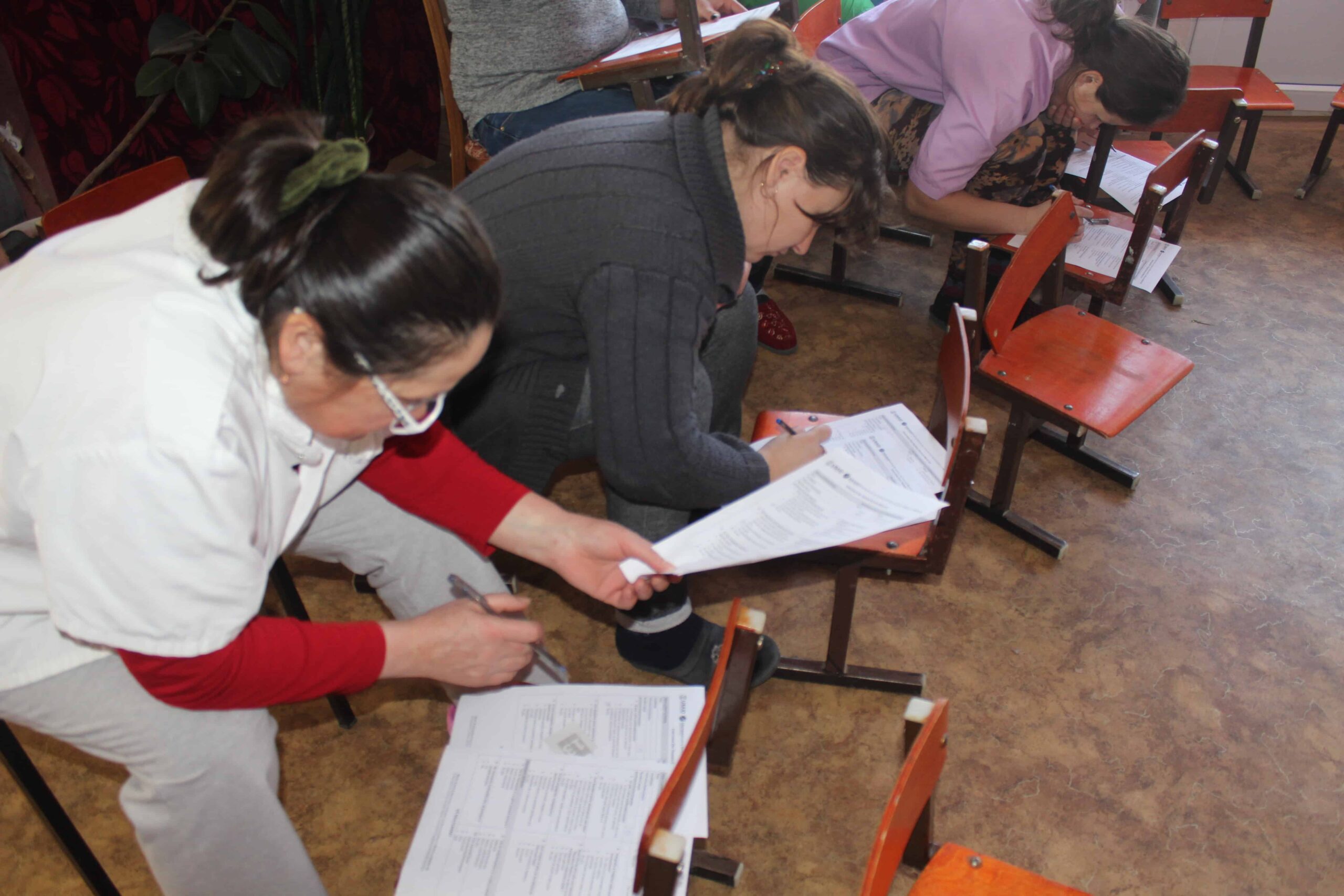 Three women sit bent over looking at printed material.