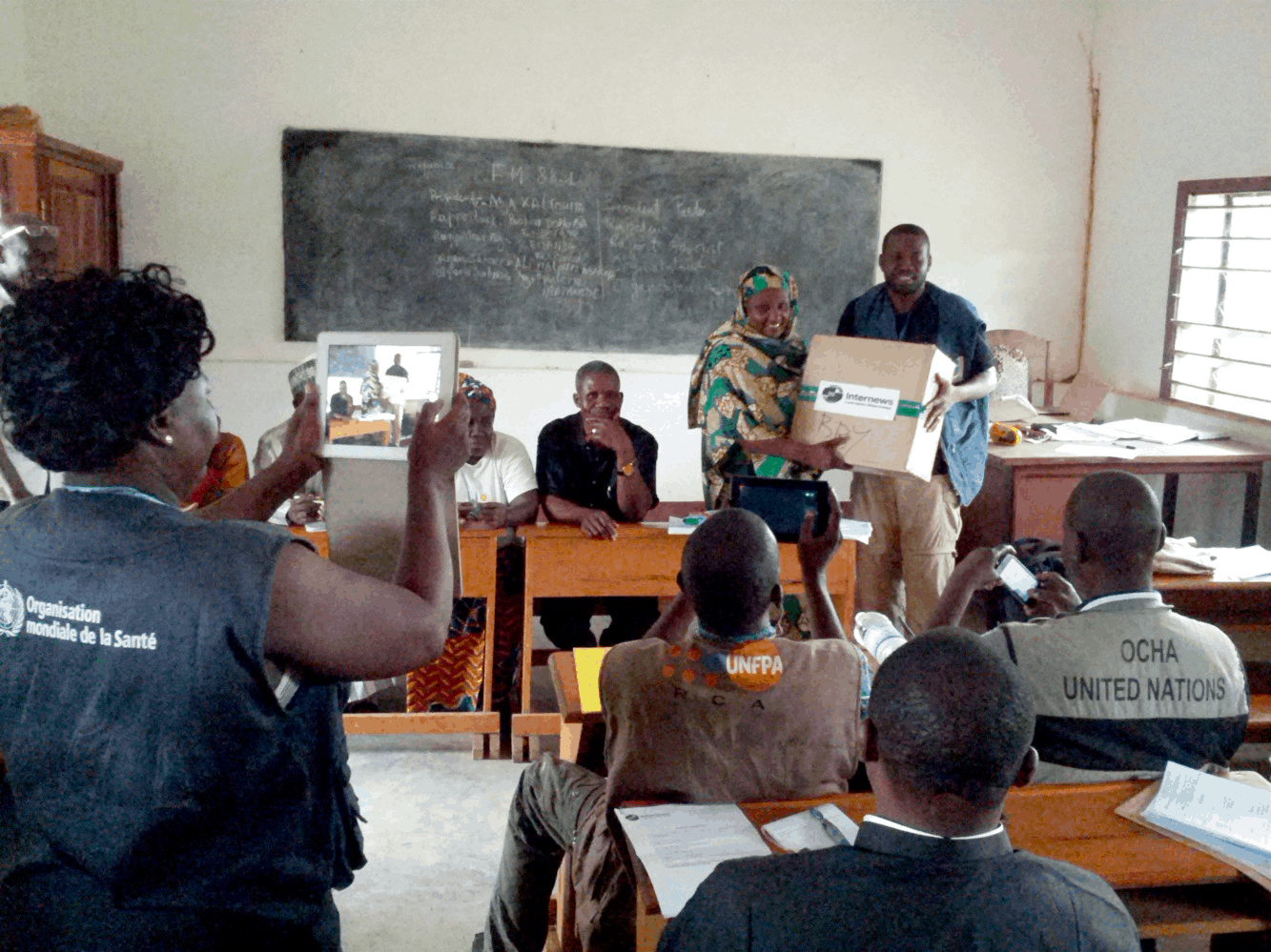 People sit in a classroom while a man hands over a box to a woman