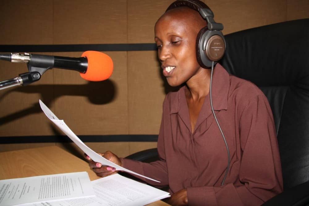 Wangari Migwi sits in the studio reading from a paper and speaking into a mic