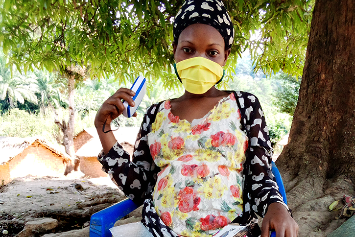 A woman sits in a chair under a tree, holding a smart phone and wearing a face mask.