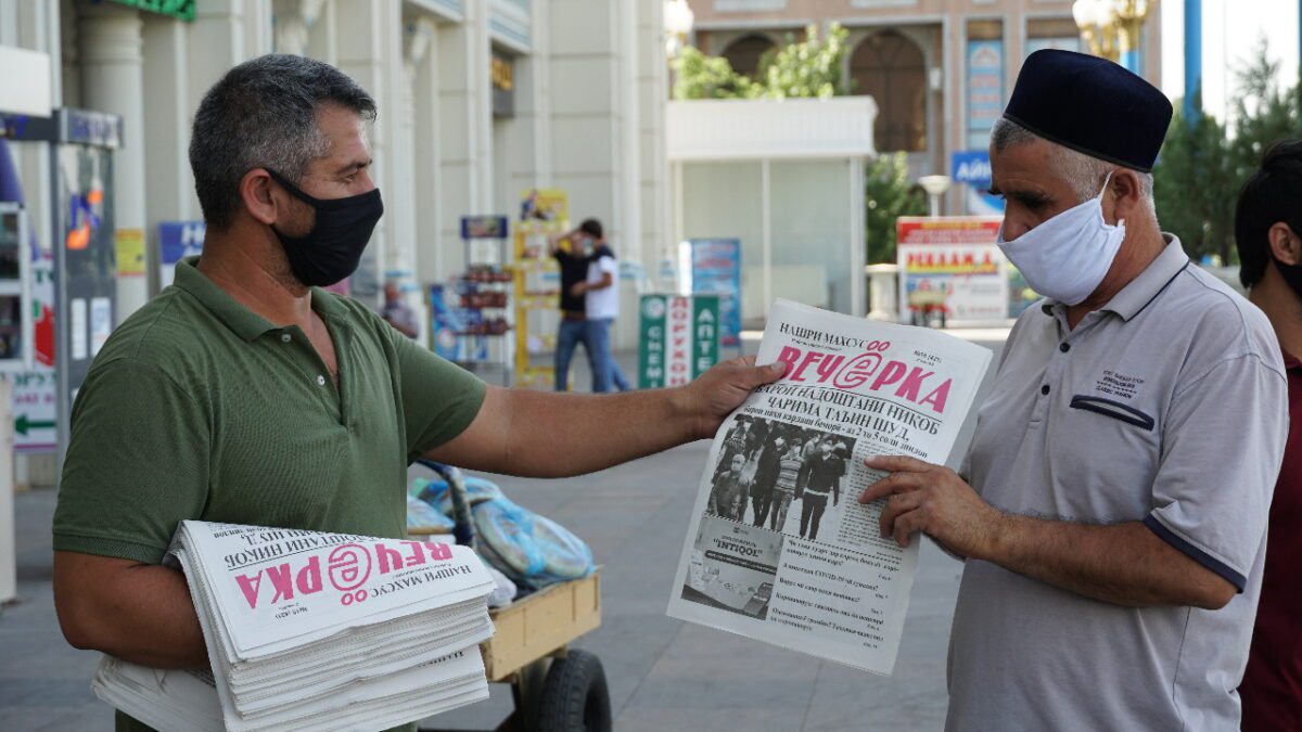 A man wearing a face covering hands a newspaper to another man