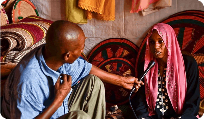 A man kneels on the floor to interview a seated woman; colorful fabrics are in the background