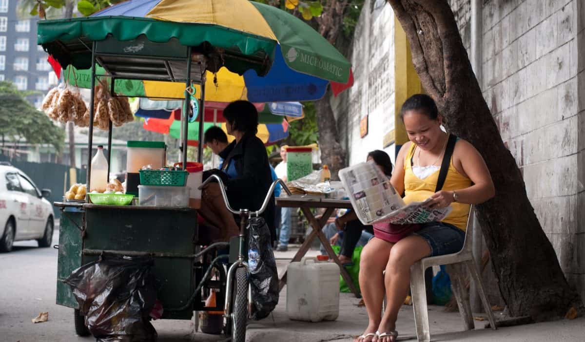 A woman sits on a chair on the street reading a newspaper; she's next to a food cart.