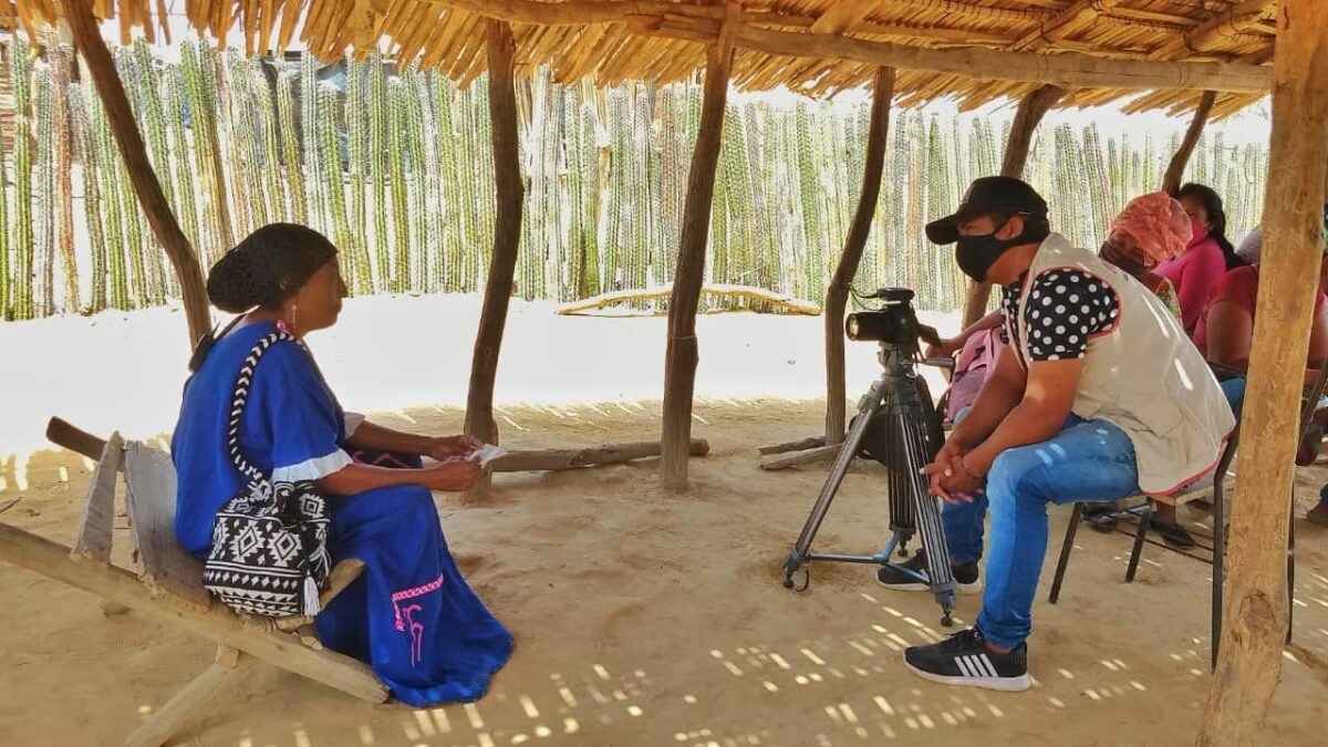 A man wearing a face mask and using a video camera sits under a tent interviewing a woman seated at a distance