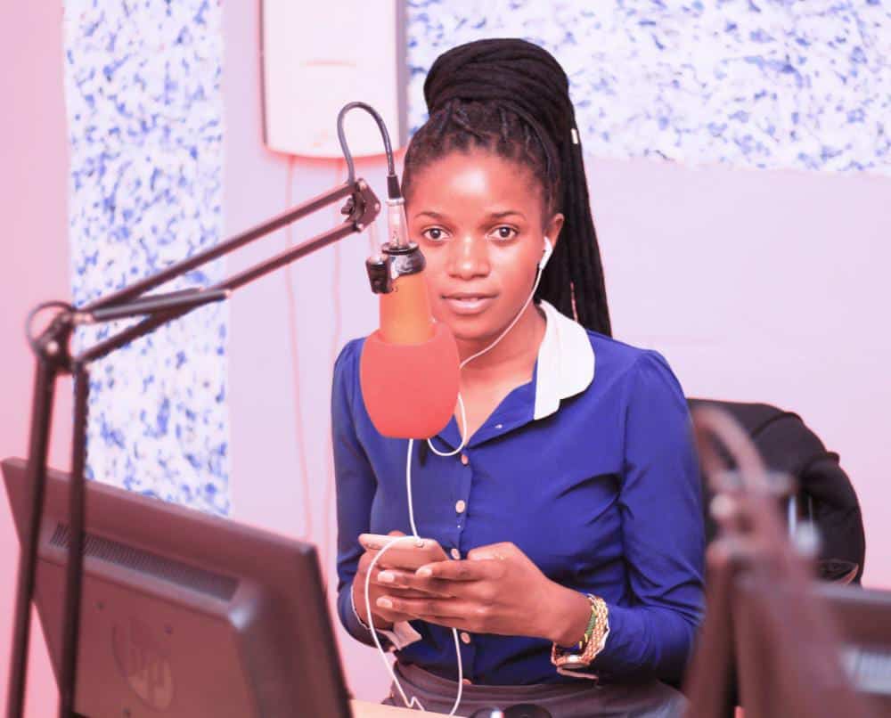 A woman stands behind a computer, speaking into a microphone.