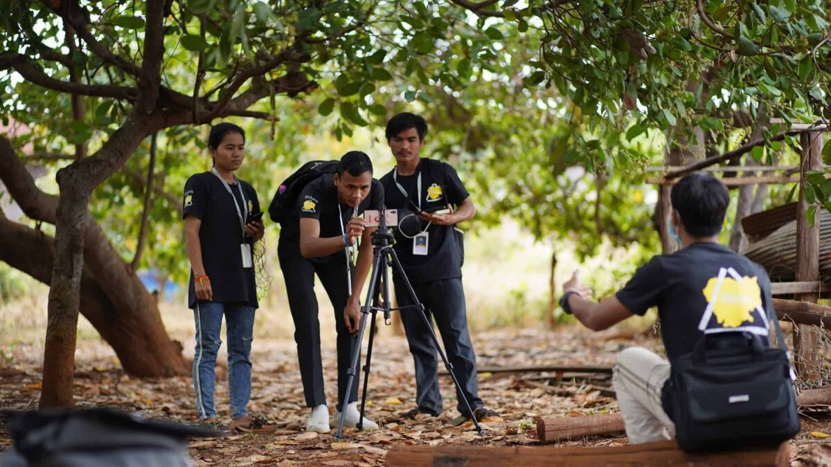 A group of young men use a video camera; they are outside under some trees