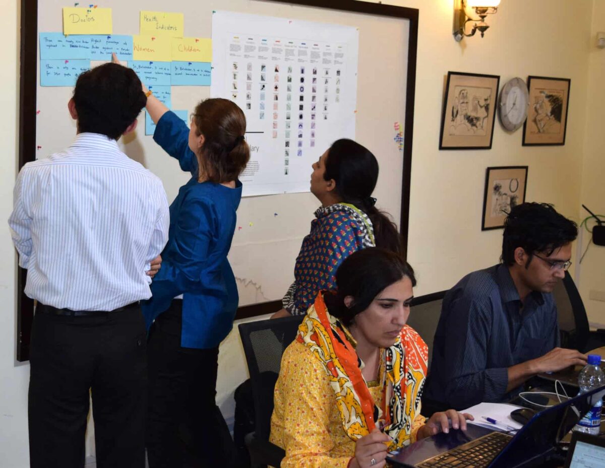 A woman writes on a sticky note on a board while a man and a woman watch; another woman and man sit at a table working on laptop computers.