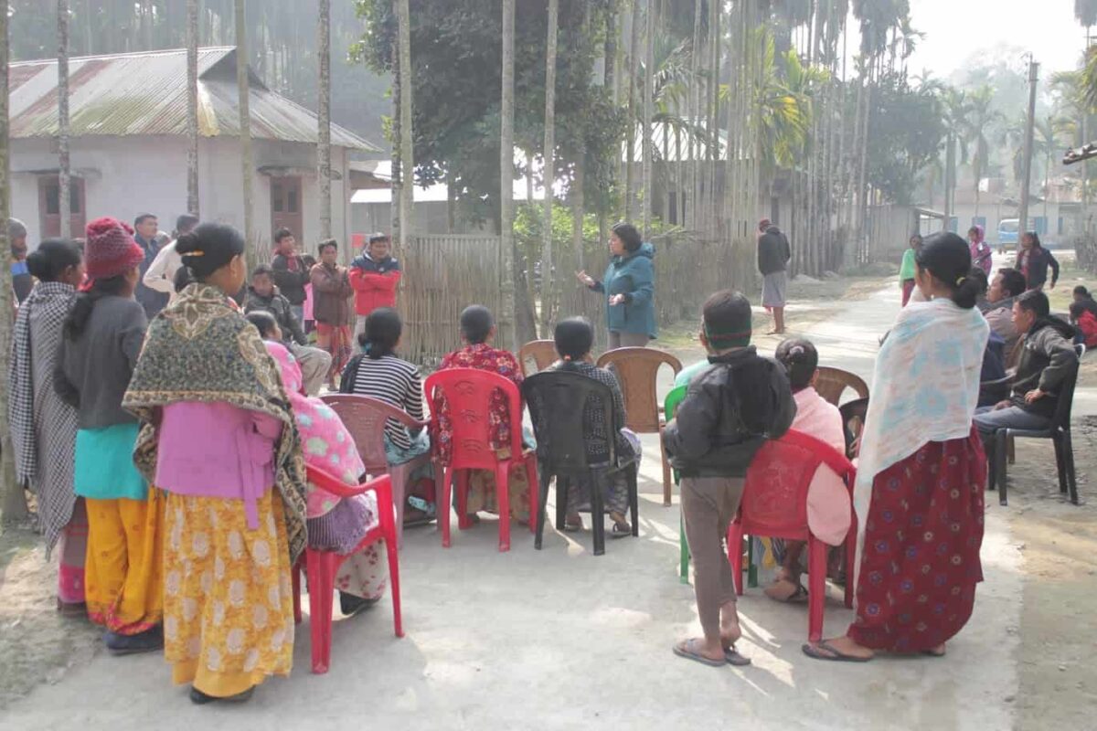 A group of people sit outside on chairs facing a woman who is standing and talking.