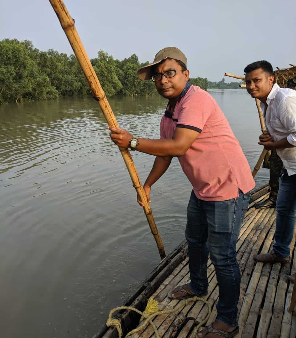 A man holds a long pole while he stands on a barge in a river.