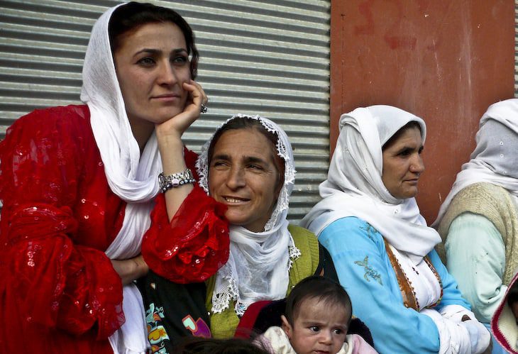 Four women sit outside a building; one holds an infant on her lap.
