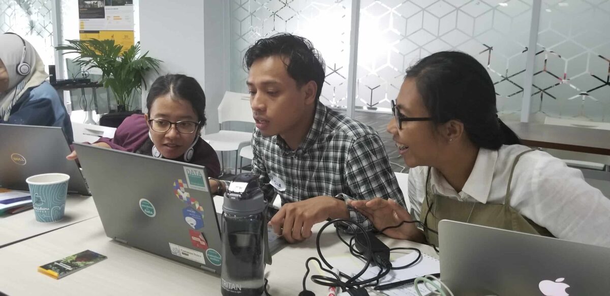 Two women and a man sit at a table, looking at a laptop computer