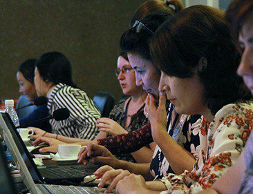 Five women sit at a table working on laptop computers.