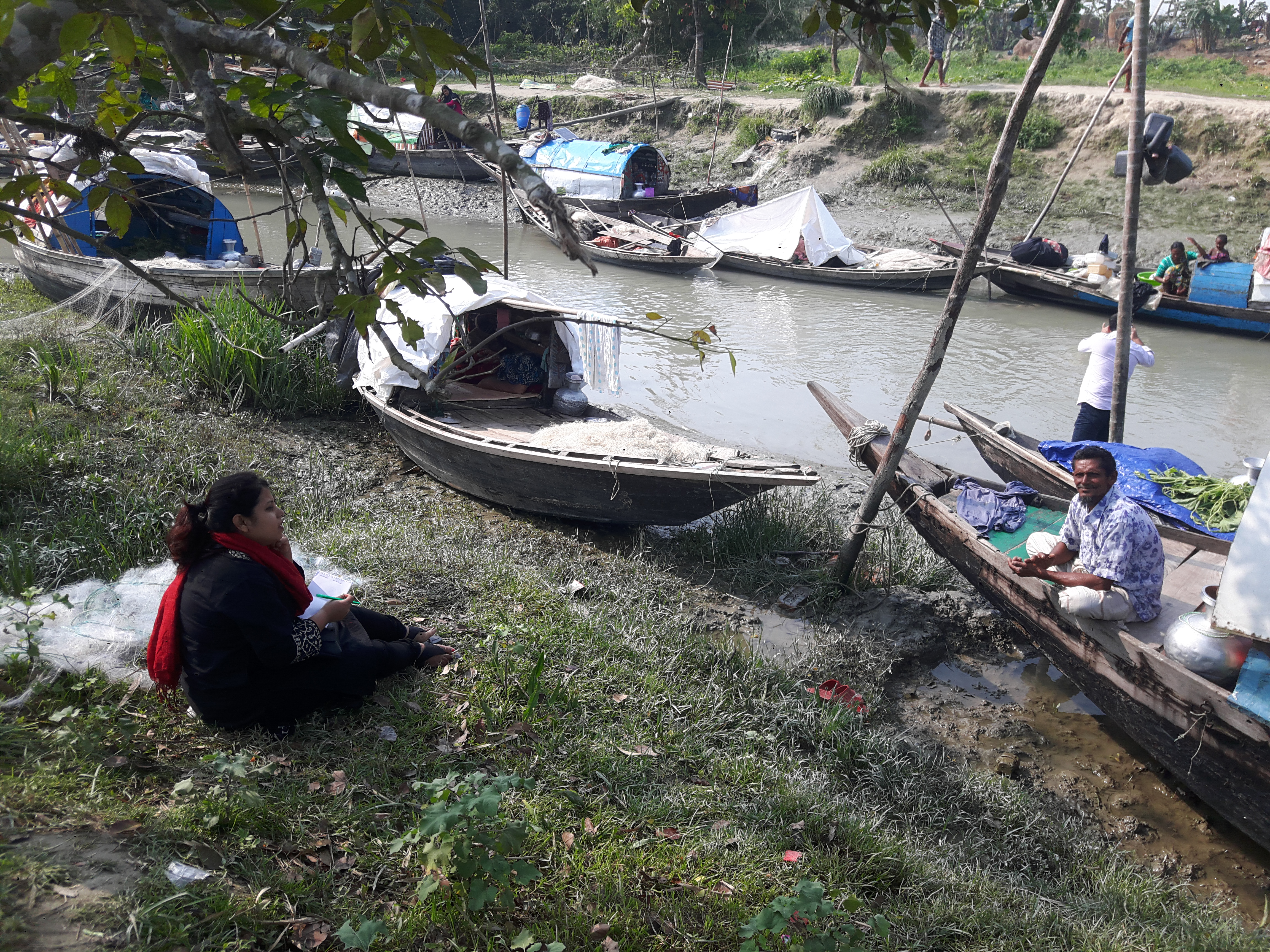 A woman sits on the banks of a river. Boats are on the shore and in the river.