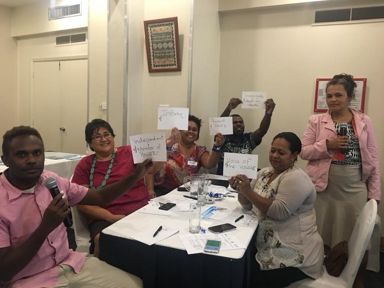 Four women and two men sit around a table holding up hand written signs