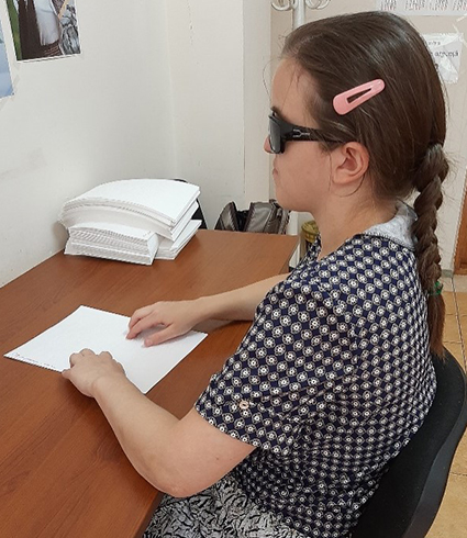 A woman wearing sunglasses sits at a table reading a Braille book