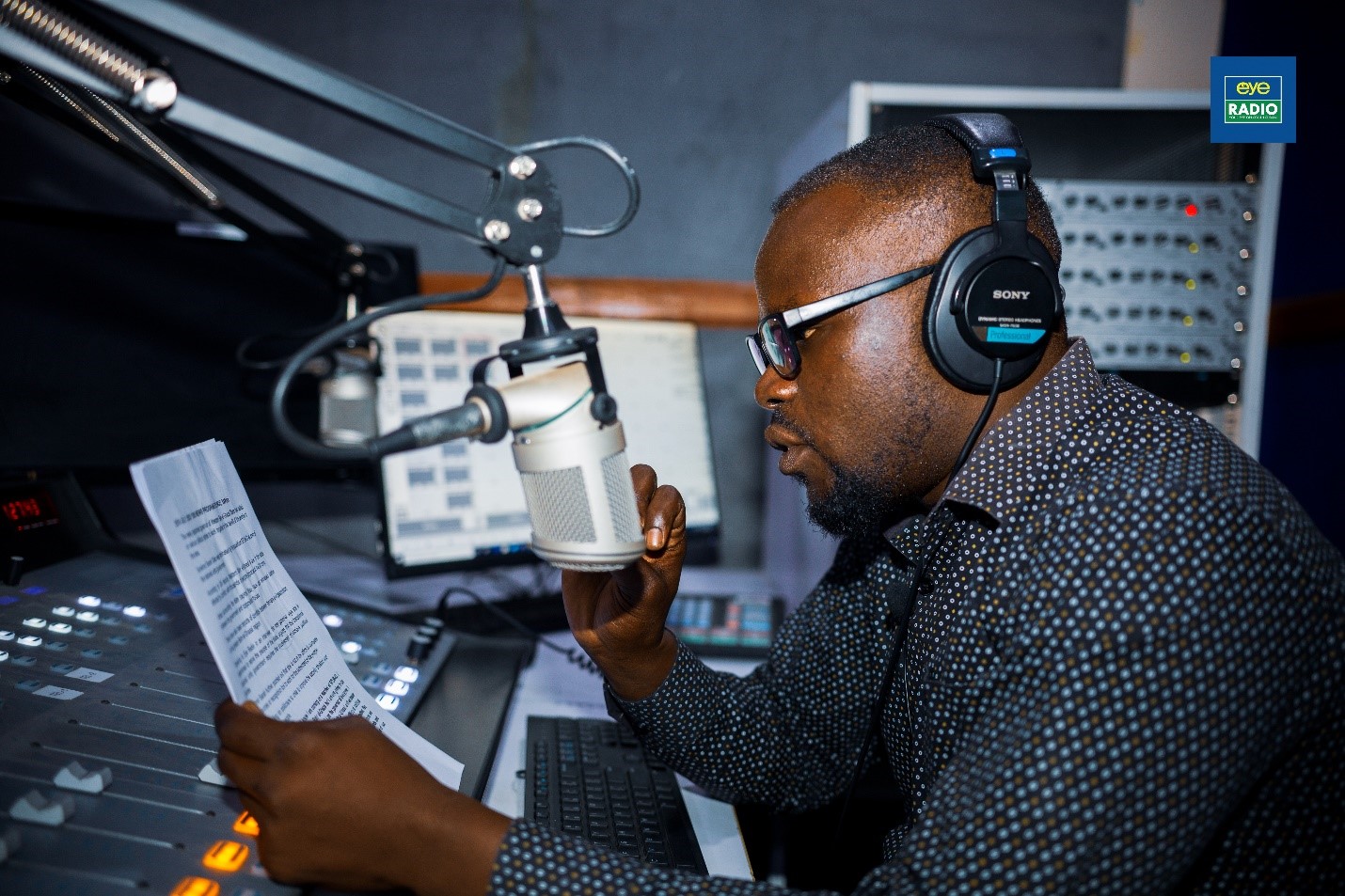 A man sits in front of a mic in a radio studio reading from a piece of paper