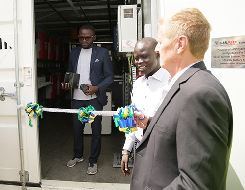 One man stands in a doorway behind a ribbon; two men stand outside, one holding scissors