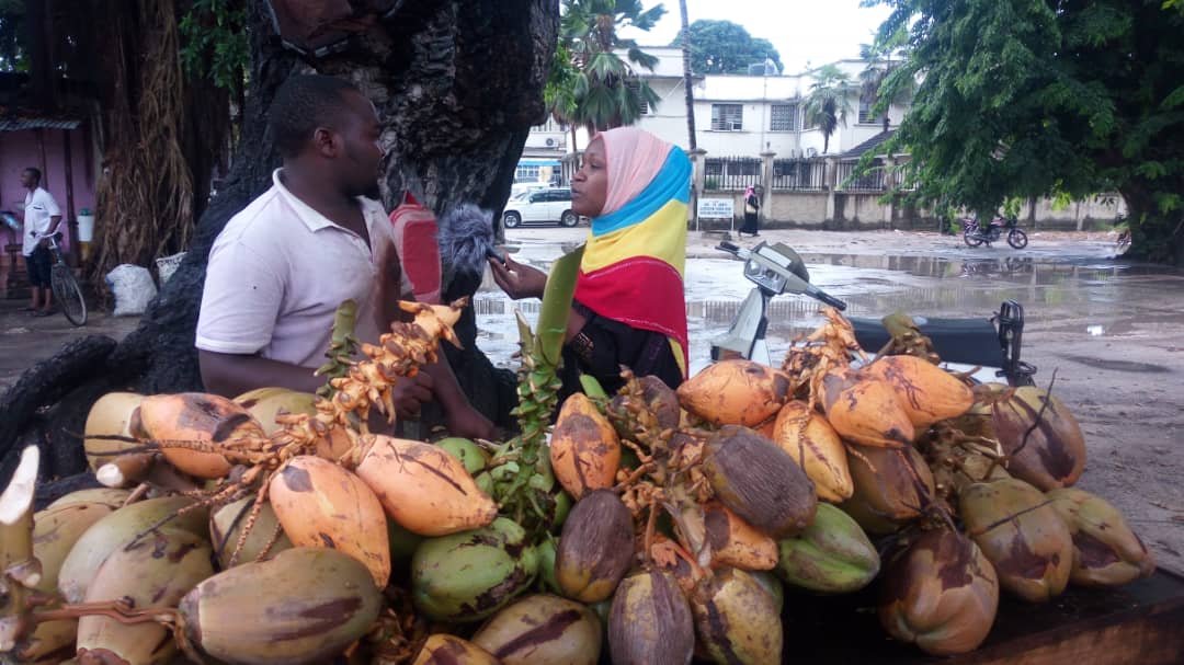 A woman holds a mic interviewing a man who is standing by a pile of coconuts.