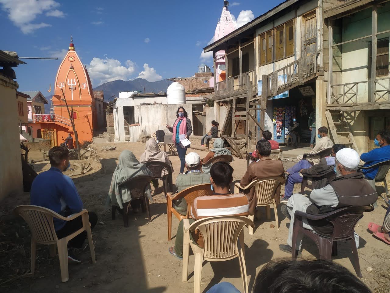 A woman stands in front of a group of people seated on plastic chairs outside some houses