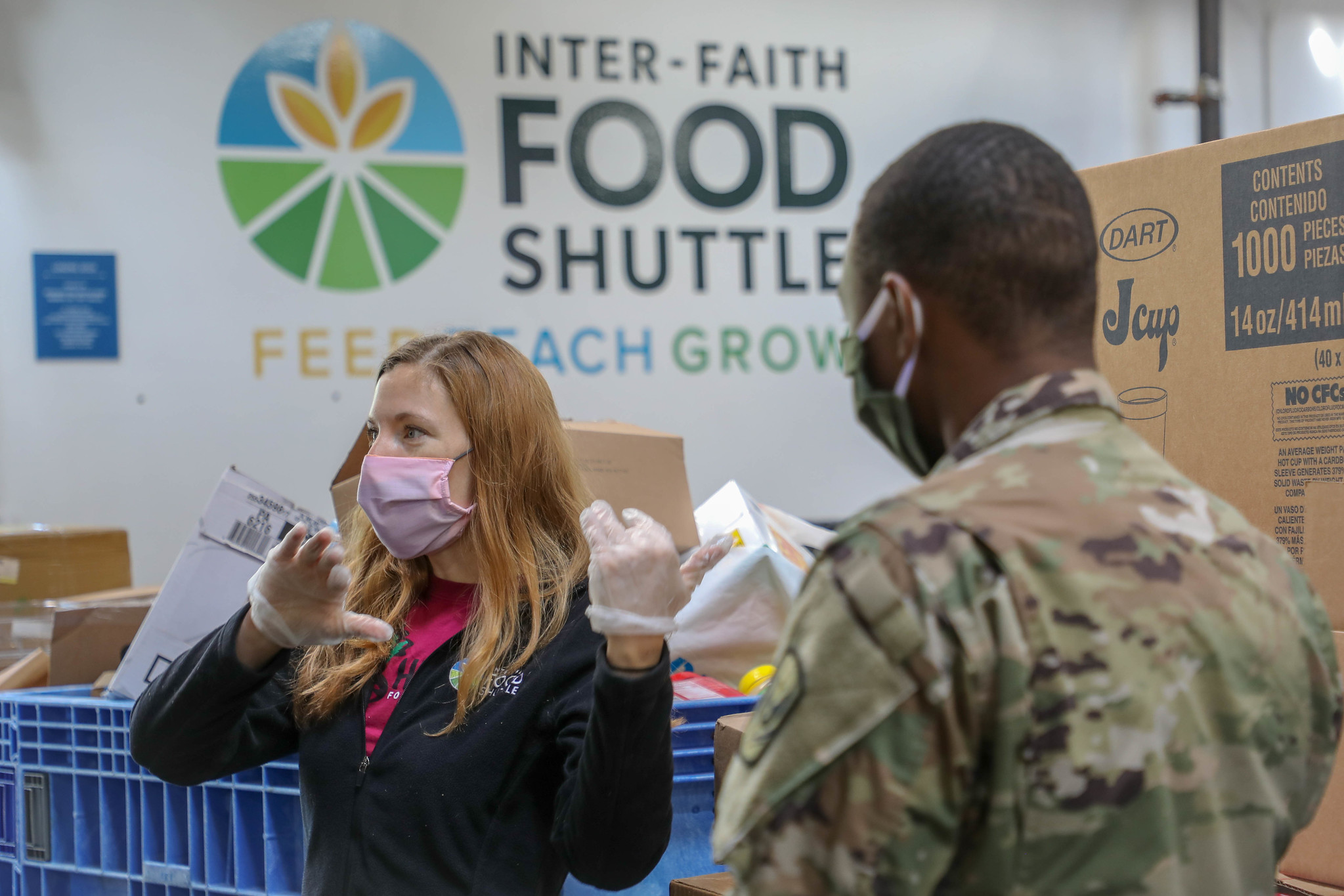 A woman wearing a mask and gloves stands next to a man wearing a mask and fatigues and in fromt of a Food bank sign and groceries.