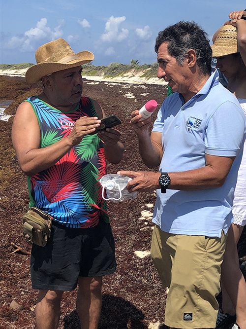 One man interviews another with a mic, standing on the beach