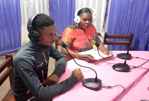 A man and a woman sit at mics in front of a table in a radio studio
