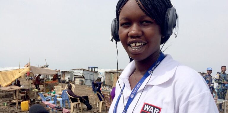 A young woman wearing headphones stands in a refugee camp.