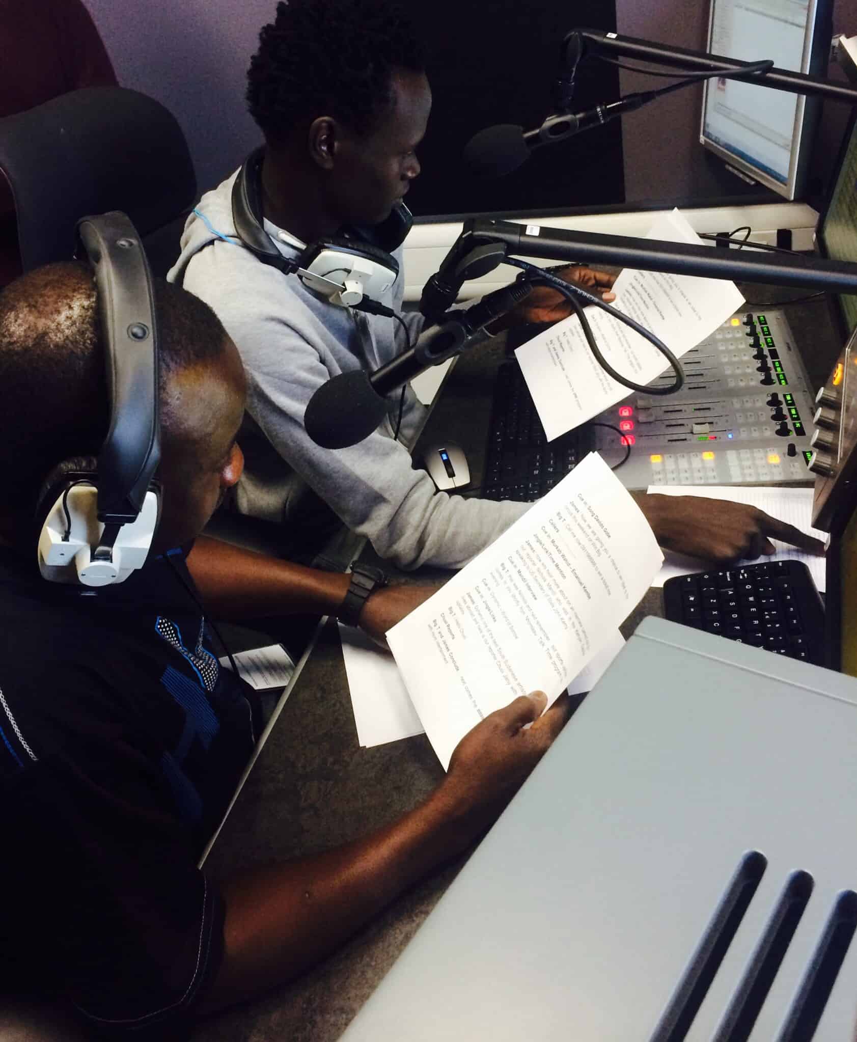 Two men sit at mics in a radio studio looking at scripts.