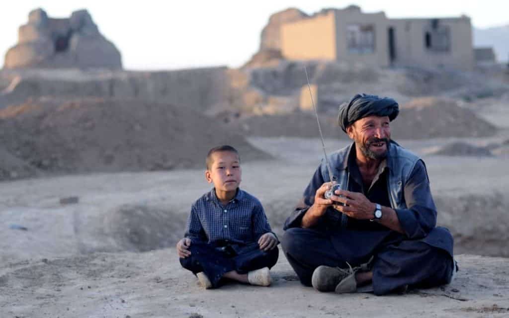 A man holding a radio and a young boy sit together on the sand - there are some brick buildings behind them