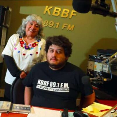 A man sits at a table with an older woman standing behind him. "KBBF 89.1 FM" is on the wall behind them.
