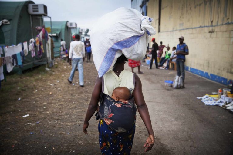 A woman carrying a baby and holding a sack on her head walks down a dirt road.